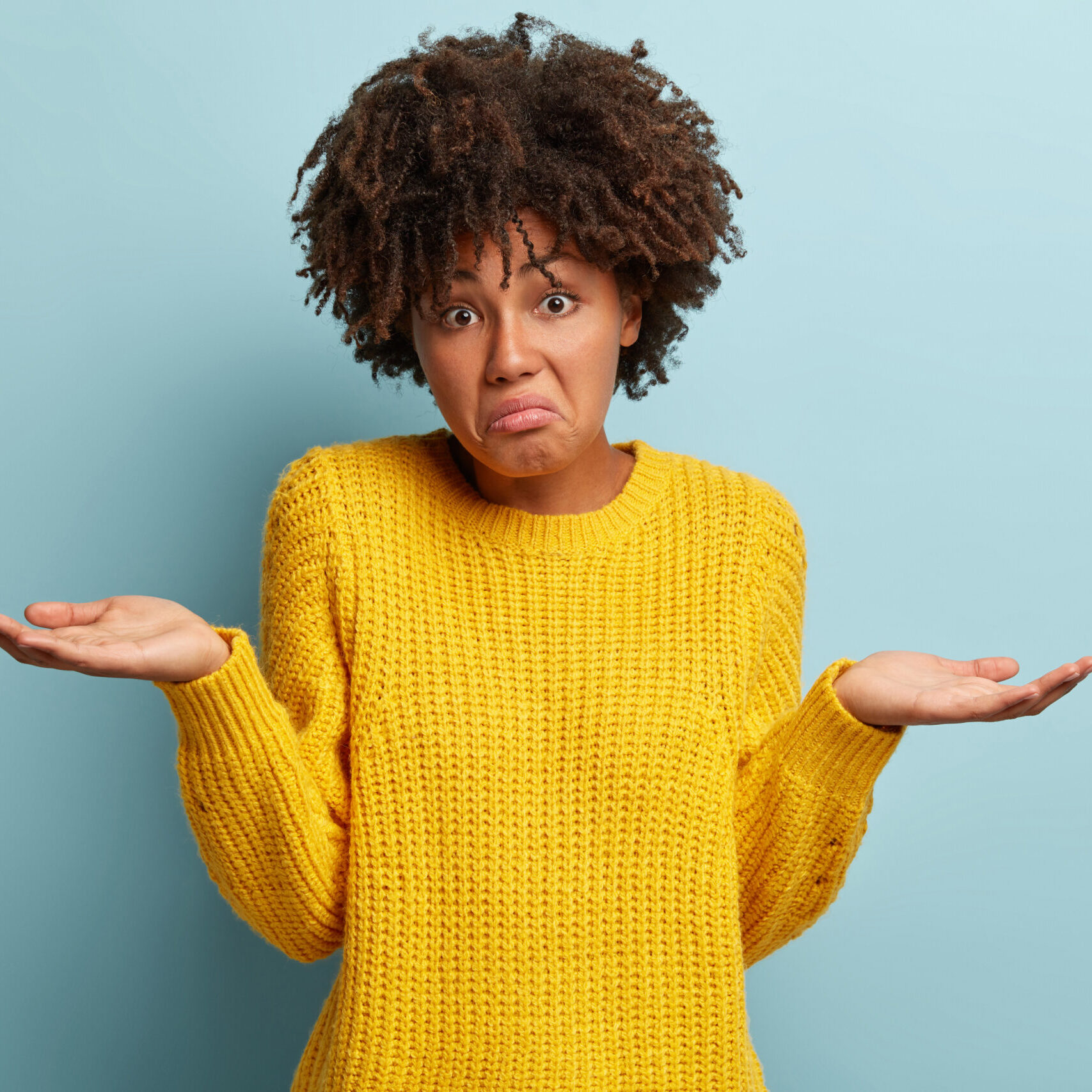 Hmm, I have no idea. Puzzled attractive curly Afro American woman spreads hands sideways, wears casual yellow loose jumper, purses lips, models over blue background, feels apathy and uncertainty